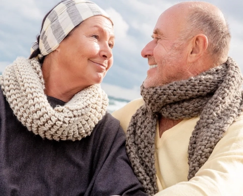 Ein älteres Ehepaar sitzt eng umschlungen im Freien und schauen sich mit liebevollen, faltigen Augen an - ihre Gesichter sind von tiefer Zuneigung und unbeschwerten Momenten geprägt. Die Frau trägt eine gemusterte Mütze und einen dicken, hellgrauen Strickschal, der Mann einen ähnlichen Schal in warmen Naturtönen. Der bewölkte Himmel im Hintergrund unterstreicht die Gemütlichkeit der Szene. Das Bild vermittelt ein Gefühl von Geborgenheit, Vertrauen und der Gewissheit, dass das Leben auch trotz Inkontinenz voller schöner, sorgenfreier Augenblicke sein kann.