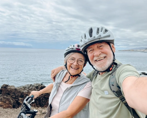Ein älteres Ehepaar macht ein fröhliches Selfie während einer gemeinsamen Fahrradtour am Meer. Beide tragen weiße Fahrradhelme und strahlen vor Lebensfreude - die Frau mit grauer Kurzhaarfrisur und Brille lächelt herzlich, während der Mann mit grauem Vollbart ausgelassen lacht. Sie umarmen sich liebevoll, im Hintergrund erstreckt sich das weite, bläuliche Meer unter einem bewölkten Himmel. Das Bild vermittelt pure Unbeschwertheit und Lebensqualität - zwei Menschen, die trotz ihres Alters aktiv und voller Energie gemeinsame Abenteuer erleben, ohne sich durch körperliche Einschränkungen davon abhalten zu lassen. Die spontane, glückliche Stimmung zeigt: Mit den richtigen Hilfsmitteln ist ein aktives, sorgenfreies Leben in jedem Alter möglich.